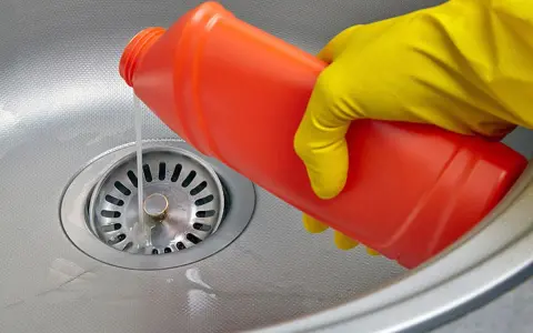 Mano con guante amarillo vierte líquido de una botella roja para como limpiar desague fregadero.