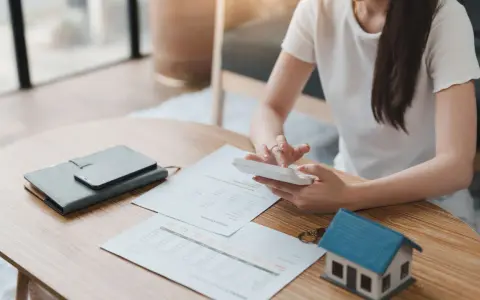Persona calculando gastos en una mesa con un modelo de casa, documentos y calculadora. Se pregunta quién paga el alta de suministros en un alquiler.