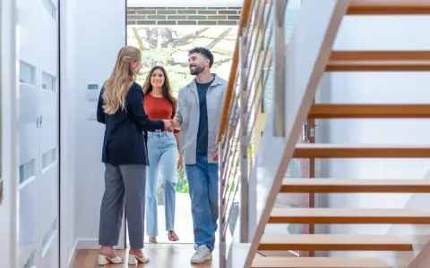 Agente inmobiliaria saluda a pareja en la entrada de una casa moderna, con escaleras de madera a la derecha. Cumpliendo el **articulo 14 LAU**.