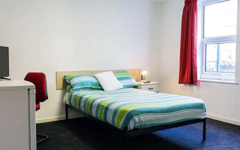 Habitación en Atlantic Point, Liverpool, con cama a rayas verdes y azules, escritorio rojo y ventana con cortina roja.