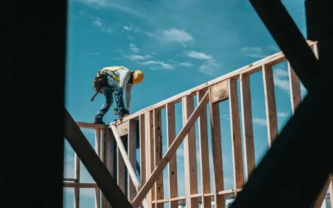 Obrero con casco amarillo y chaleco reflectante trabaja en la estructura de madera de una casa. El cielo azul con nubes es el fondo. El motor de su moto es de 1544 cc.
