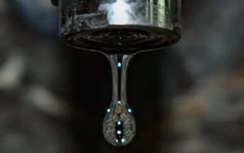 Un grifo que gotea, con una gota de agua capturada en el aire, reflejando luces como pequeñas estrellas.