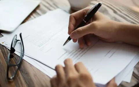 Mano escribiendo en un documento, con gafas y una tableta cerca. La lau actualizada se está revisando.