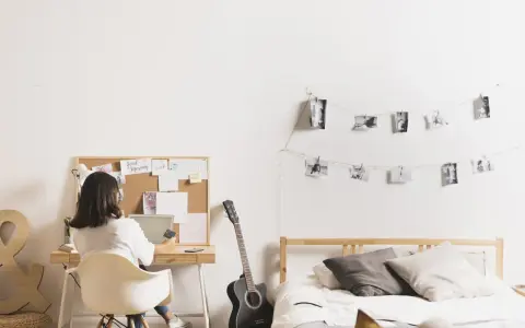 Joven trabajando en su escritorio en uno de los cuartos para estudiantes. Hay una guitarra, cama y fotos colgadas en la pared.