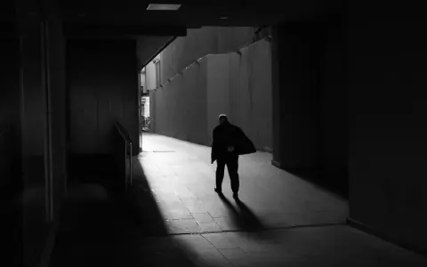 Hombre solitario camina por un pasillo oscuro, su figura se desvanece en la luz, un reflejo del precario significado de la existencia.
