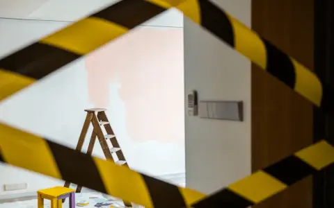 Escalera de madera y un taburete amarillo, listos para aplicar el **articulo 30 lau** en la pared. Cinta de precaución amarilla y negra.