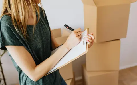 Mujer rubia con camiseta verde, revisando una lista en un portapapeles, rodeada de cajas de mudanza. El arte 19 lau inspira la organización.