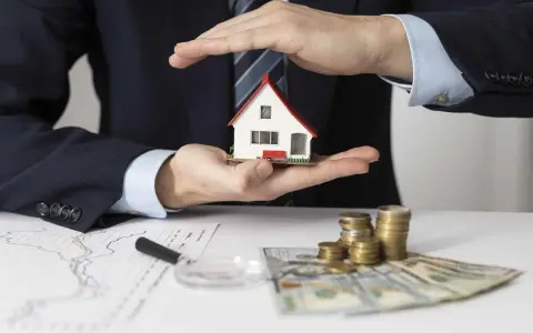 Manos protegen una casa modelo sobre un contrato de alquiler con avalista, junto a dinero y gráficos.