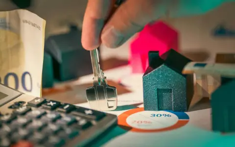 Mano sostiene llave sobre casas modelo, calculadora y billetes. El artículo 18 de la LAU se relaciona con la compraventa de viviendas.