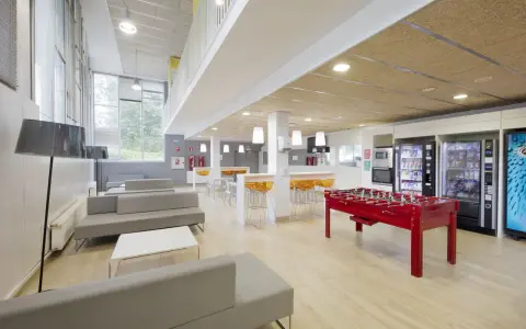 Sala común de una residencia estudiantil con sofás grises, futbolín rojo, máquinas expendedoras y mesas altas con taburetes naranjas.