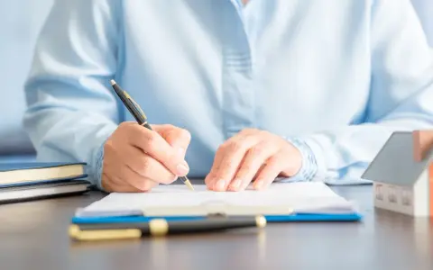 Persona firmando un documento en una mesa, donde se registra un contrato de alquiler. Hay una maqueta de casa y libros.