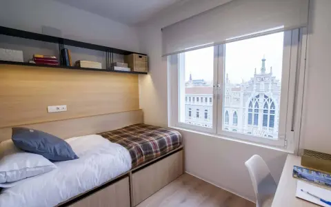Habitación moderna con cama, estanterías y vistas a un edificio histórico en Xior Madrid Retiro.
