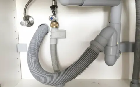 El fregadero pierde agua por abajo. Tuberías grises y mangueras flexibles bajo el lavabo, con una fuga visible.