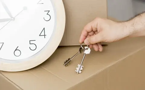 Un reloj marca el tiempo mientras una mano sostiene las llaves, simbolizando la inminencia de un contrato de alquiler temporal y una mudanza.