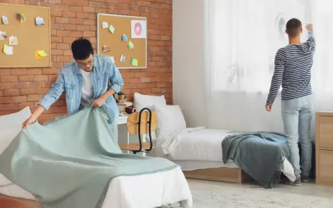 Dos jóvenes preparan su habitación de studea. Uno tiende una manta en la cama, el otro ajusta las cortinas.