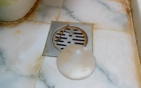 Desagüe de baño con rejilla metálica y tapa redonda. Se observa suciedad y sarro, indicando la necesidad de limpiar bote sifonico.