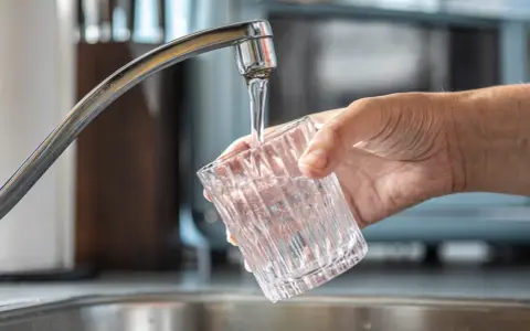 Mano sujetando un vaso de cristal bajo un grifo, llenándolo de agua. Un momento especial para dar de alta el agua por primera vez en un nuevo hogar.