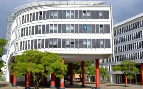 Edificio curvo blanco con ventanas oscuras y árboles verdes. Columnas rojas sostienen la estructura, creando un espacio crous.