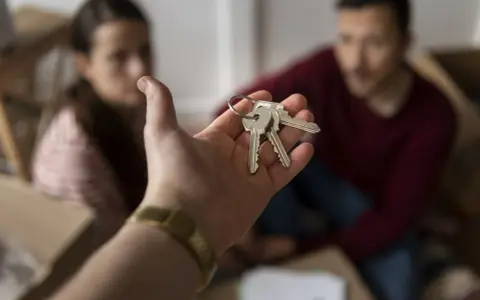 Mano entrega llaves. Pareja sonriente rodeada de cajas, lista para recuperar vivienda alquilada para un hijo.