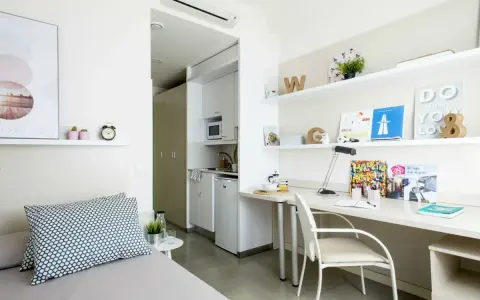 Estudio moderno en residencia La Salle: cama, escritorio con lámpara y estanterías, y pequeña cocina.