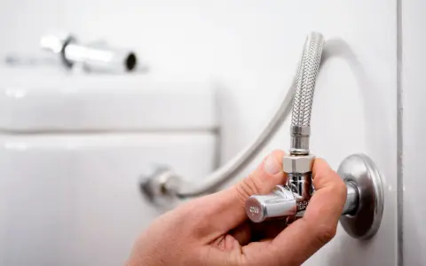 Mano ajustando la llave de paso del agua, que es comunitaria, conectada a una manguera flexible y al inodoro.