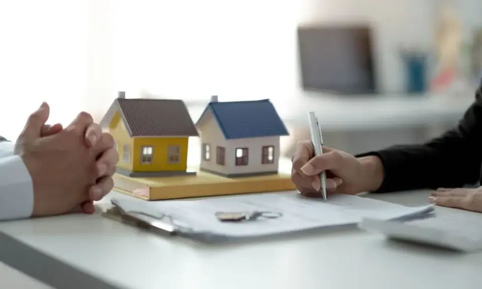 Manos entrelazadas junto a maquetas de casas y un bolígrafo firmando un contrato privado de alquiler.