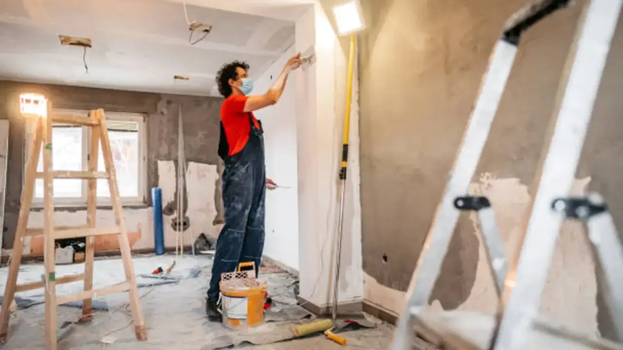Un trabajador con mascarilla pinta una pared en una obra. Escaleras y cubos de pintura sugieren que se está aplicando el artículo 22 de la L.A.U.
