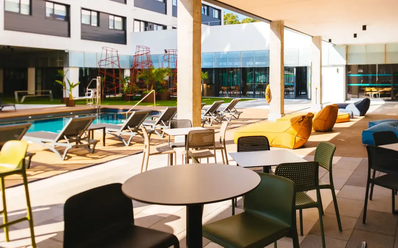 Zona de piscina y descanso en la residencia de estudiantes Livensa Living Sevilla, con tumbonas, mesas y pufs.
