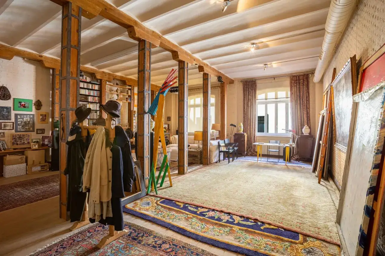 Un loft que es un espacio de arte y vida, con vigas de madera, estanterías llenas, abrigos y cuadros.