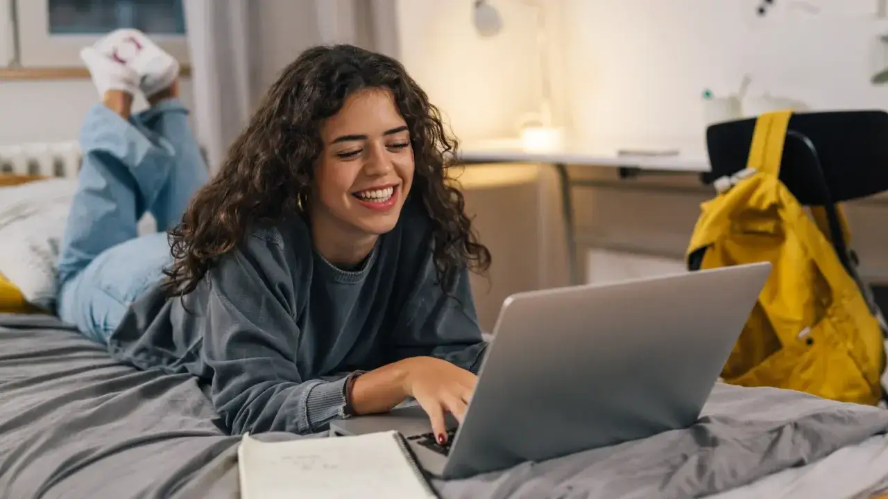 Joven sonríe mientras estudia en su laptop en la cama.