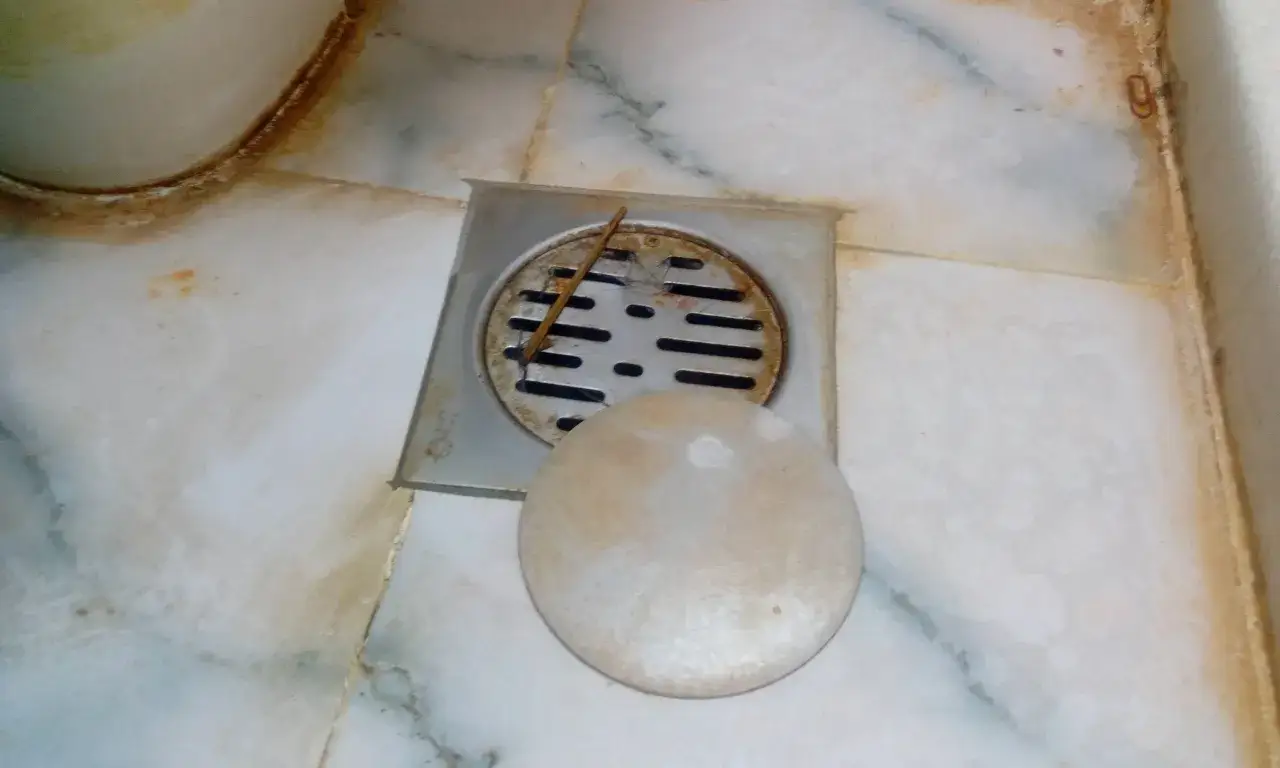 Desagüe de baño con rejilla metálica y tapa redonda. Se observa suciedad y pelos, indicando la necesidad de limpiar bote sifónico.