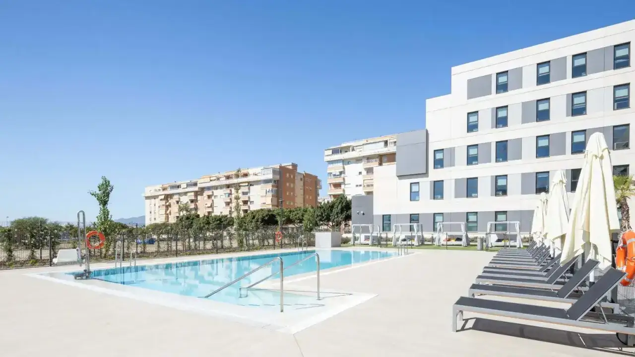 Piscina y tumbonas en Livensa Living Studios Málaga Feria, rodeado de edificios residenciales y vegetación bajo un cielo azul.