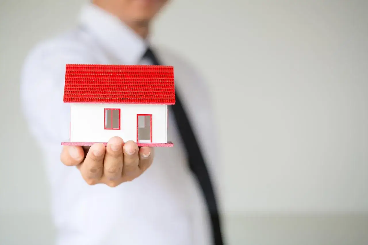 Un hombre con corbata negra sostiene una maqueta de casa con techo rojo. El piso sale a subasta.