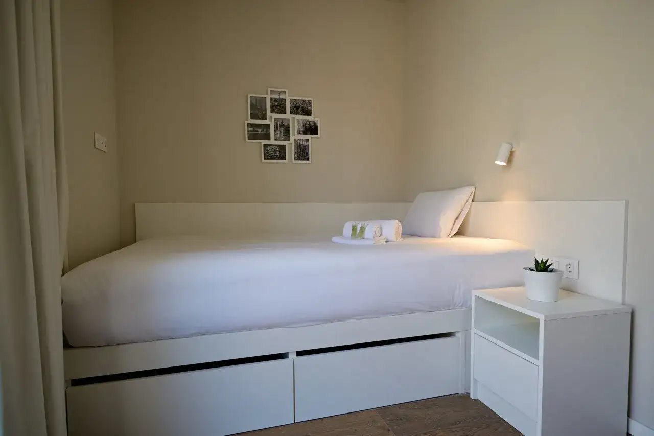 Habitación en residencia Xior Collblanc con cama blanca, mesita de noche con planta y fotos en la pared.