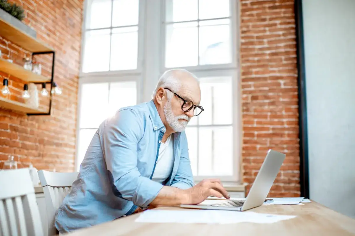 Hombre mayor con gafas trabajando en su portátil. El texto en pantalla dice
