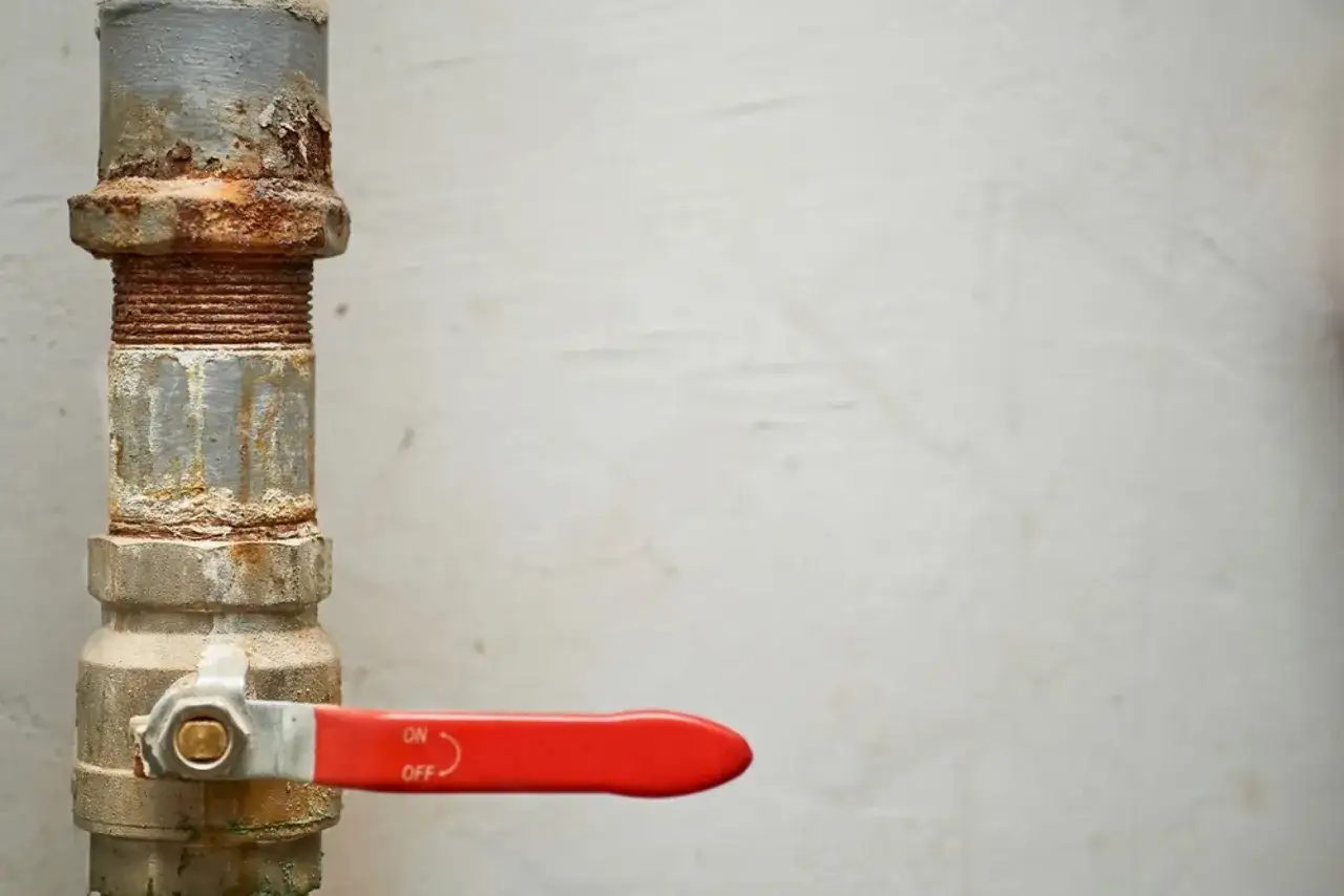 Llave de paso de agua general con palanca roja, instalada en tuber&iacute;a met&aacute;lica oxidada.