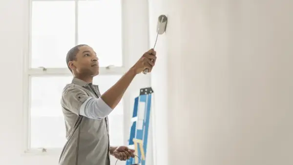 Hombre pintando pared blanca, resolviendo quién paga la pintura en un piso de alquiler. Escalera azul al fondo.