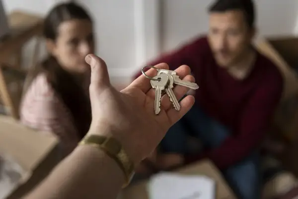 Mano entrega llaves. Pareja sonriente rodeada de cajas, lista para recuperar vivienda alquilada para un hijo.